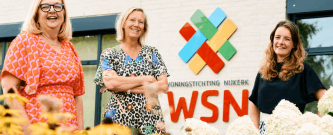 Drie vrouwen staan voor het kantoor van Woningstichting Nijkerk bij het WSN-logo, met op de voorgrond gele bloemen.