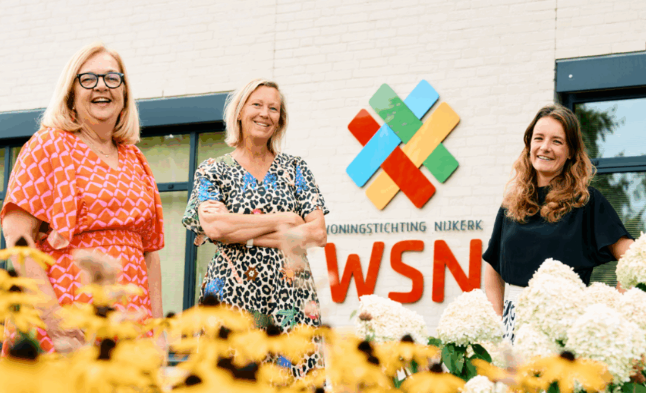 Drie vrouwen staan voor het kantoor van Woningstichting Nijkerk bij het WSN-logo, met op de voorgrond gele bloemen.