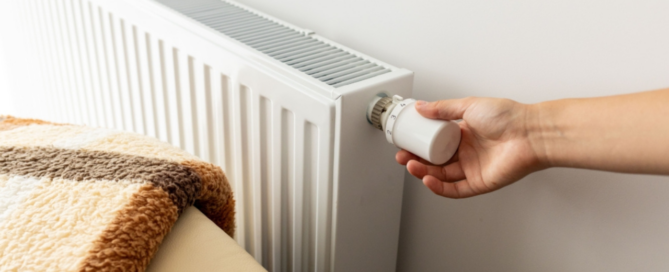 Hand draait aan de thermostaatknop van een witte radiator naast een bed met een warme deken