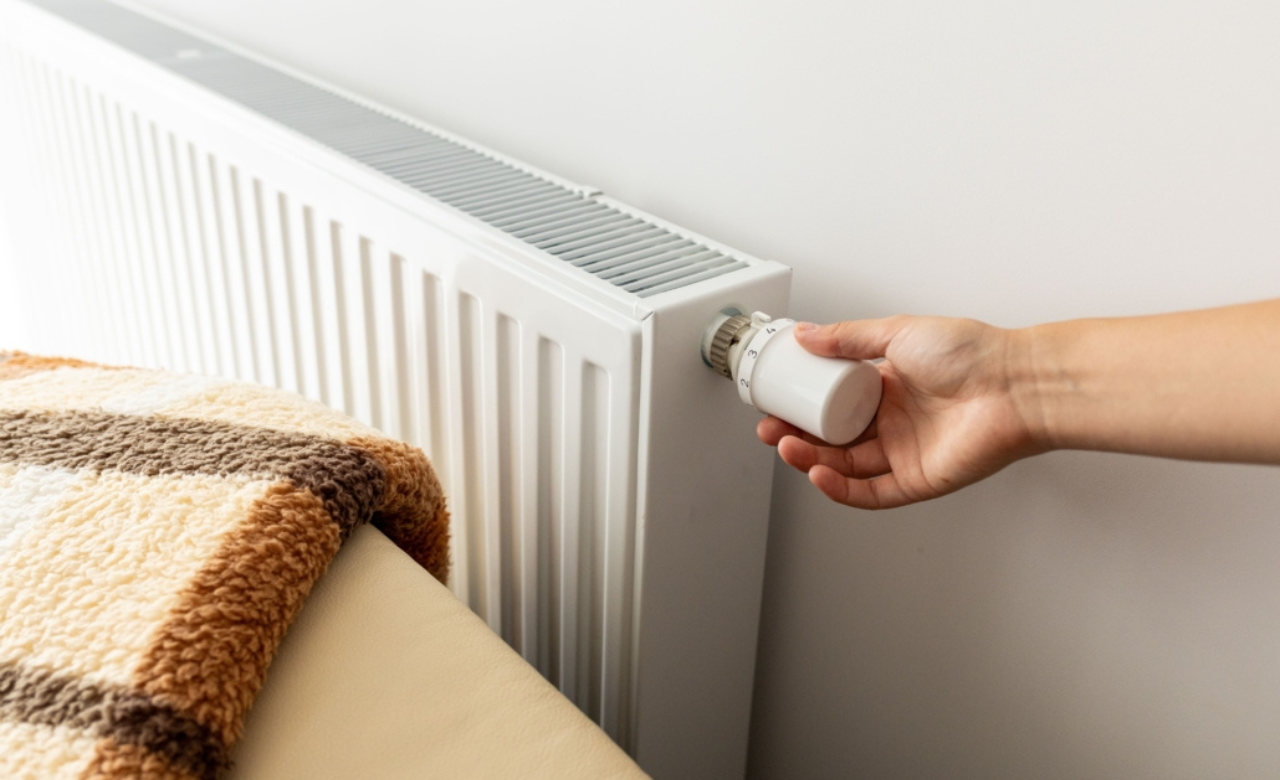 Hand draait aan de thermostaatknop van een witte radiator naast een bed met een warme deken