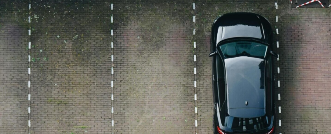 Luchtfoto van een half lege parkeerplaats met een zwarte auto naast groenbegroeiing.