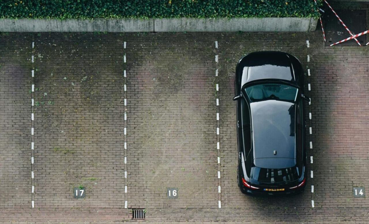 Luchtfoto van een half lege parkeerplaats met een zwarte auto naast groenbegroeiing.