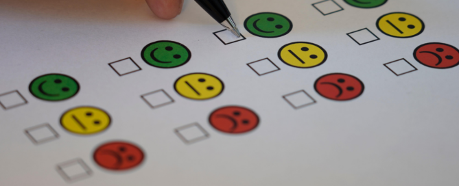 Formulier met rijen gekleurde smileys van groen blij tot rood verdrietig met lege vakjes ervoor, een hand houdt een pen boven een vakje om een keuze te maken