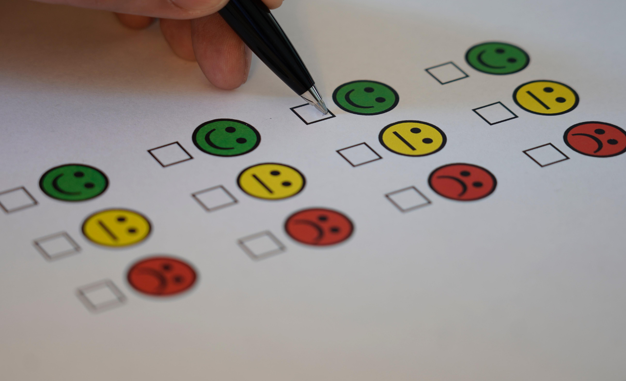 Formulier met rijen gekleurde smileys van groen blij tot rood verdrietig met lege vakjes ervoor, een hand houdt een pen boven een vakje om een keuze te maken