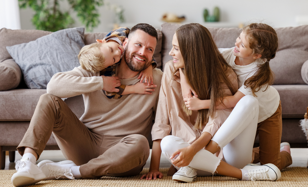 Gezin zit lachend samen op de vloer in een woonkamer en geniet van een moment thuis